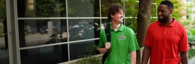 Two smiling coworkers walking outside an office building, one wearing a green Interstate Batteries polo shirt and the other in a red polo.