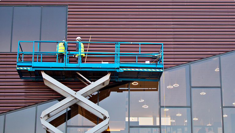 Two construction workers wearing safety vests stand on a blue scissor lift while working on the exterior of a modern commercial building with glass windows and metal panels
