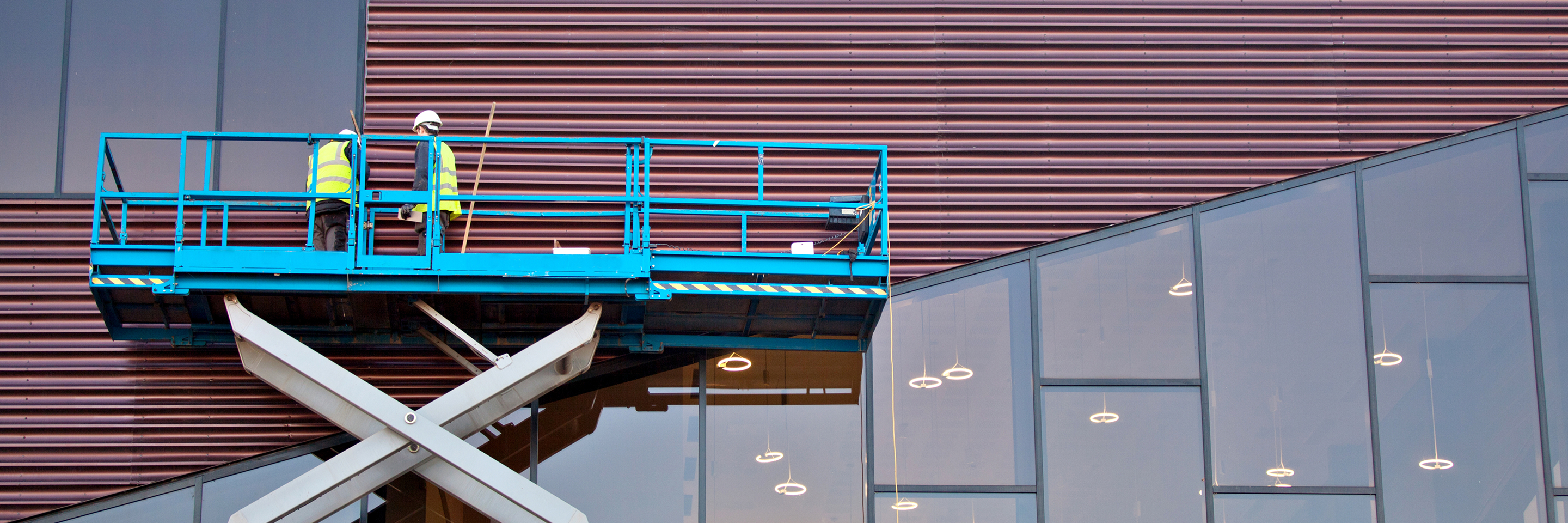 Two construction workers wearing safety vests stand on a blue scissor lift while working on the exterior of a modern commercial building with glass windows and metal panels